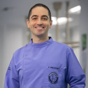 La imagen muestra a una persona de frente en un entorno interior tipo clínico o académico. Viste un uniforme médico de color violeta con botones laterales y un logotipo institucional bordado en el pecho. El fondo es desenfocado, con iluminación blanca y elementos propios de un espacio de atención en salud.