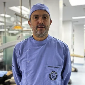La imagen muestra a una persona de frente en un entorno clínico interior. Viste uniforme médico de color azul claro y un gorro quirúrgico del mismo tono. El fondo es un espacio de atención en salud desenfocado, con iluminación blanca y equipamiento clínico visible de manera general.