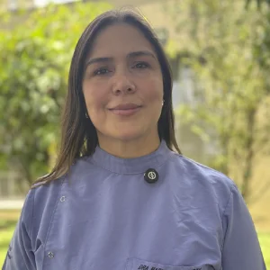 La imagen muestra a una persona de frente en un entorno exterior con vegetación verde desenfocada al fondo. Viste un uniforme clínico de color violeta claro con cuello cerrado y broches visibles. El cabello es lacio, de longitud media, suelto sobre los hombros. La iluminación es natural y el encuadre es de primer plano.