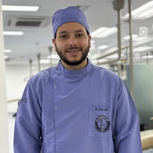 La imagen muestra a una persona de frente en un entorno clínico interior. Viste un uniforme médico de color violeta claro con botones laterales y un gorro quirúrgico del mismo tono. El fondo está desenfocado e incluye iluminación blanca y elementos propios de una sala de atención en salud.