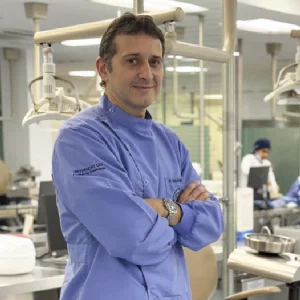 La imagen muestra a una persona en un entorno clínico interior, vistiendo un uniforme médico de color azul claro. Se encuentra de pie con los brazos cruzados. Al fondo se observan equipos odontológicos, lámparas y mobiliario propio de un espacio de atención en salud, con iluminación blanca y ambiente profesional.