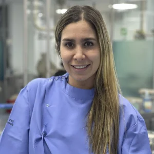 La imagen muestra a una persona de frente en un entorno clínico interior. Viste un uniforme médico de color violeta claro con botones laterales. El fondo está desenfocado y presenta equipos y mobiliario propios de un espacio de atención en salud, con iluminación blanca y ambiente profesional.