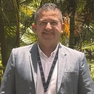La imagen muestra a una persona de frente en un entorno exterior con palmeras y vegetación al fondo. Viste un saco gris claro sobre una camisa blanca y lleva un portacarné oscuro colgado al cuello con texto repetido. La iluminación es natural y el encuadre es de medio cuerpo.