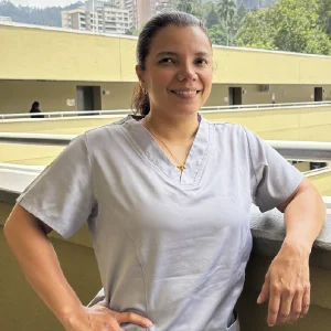 La imagen muestra a una persona en un entorno exterior institucional, apoyada sobre una baranda de concreto. Viste un uniforme clínico gris claro de manga corta y lleva un collar delgado. El fondo incluye corredores de un edificio de varios niveles y vegetación, con iluminación natural durante el día.