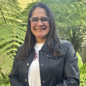 La imagen muestra a una persona de frente en un entorno exterior con abundante vegetación, principalmente hojas grandes y verdes, al fondo. Viste una chaqueta oscura abotonada sobre una camiseta clara con un diseño discreto y lleva gafas de montura clara. El cabello es largo y suelto, y la iluminación es natural con encuadre de medio cuerpo.