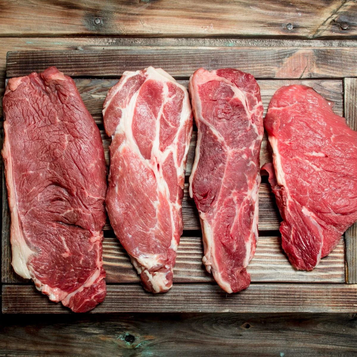 Cuatro cortes de carne cruda dispuestos en fila sobre una tabla de madera, vistos desde arriba, en un fondo rústico de madera.