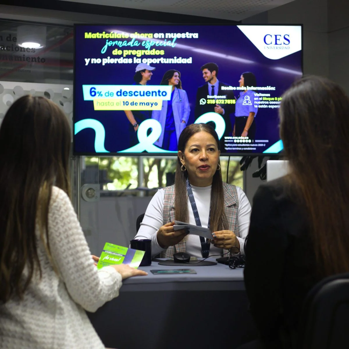 Persona atendiendo a dos visitantes en un módulo informativo de la Universidad CES. Al fondo, una pantalla promociona una jornada especial de matrículas de pregrados con 6 % de descuento hasta el 10 de mayo, e incluye logotipo institucional y datos de contacto.