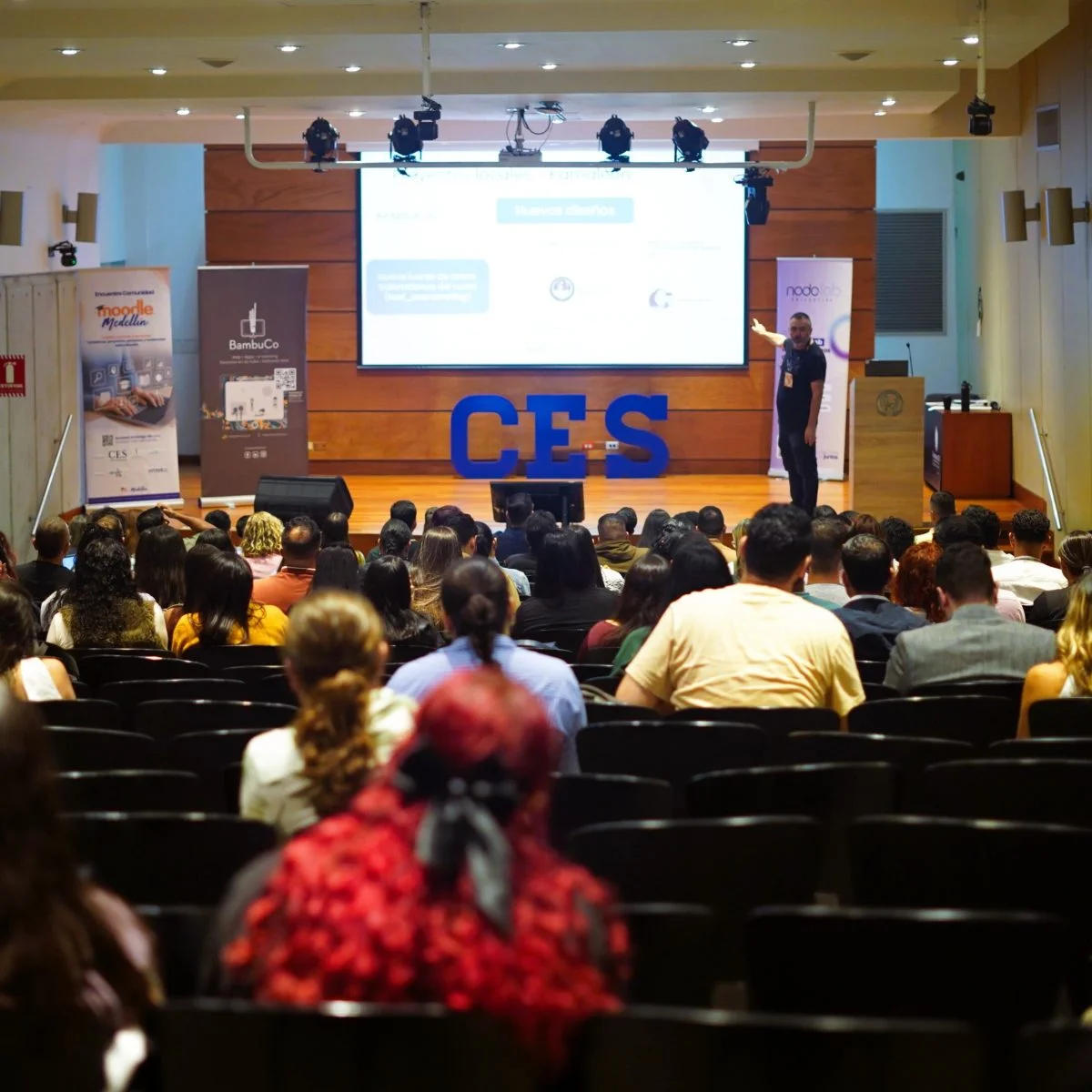 Auditorio lleno de personas sentadas frente a un escenario, donde una persona expone junto a una pantalla de proyección con diapositivas y logotipos institucionales.