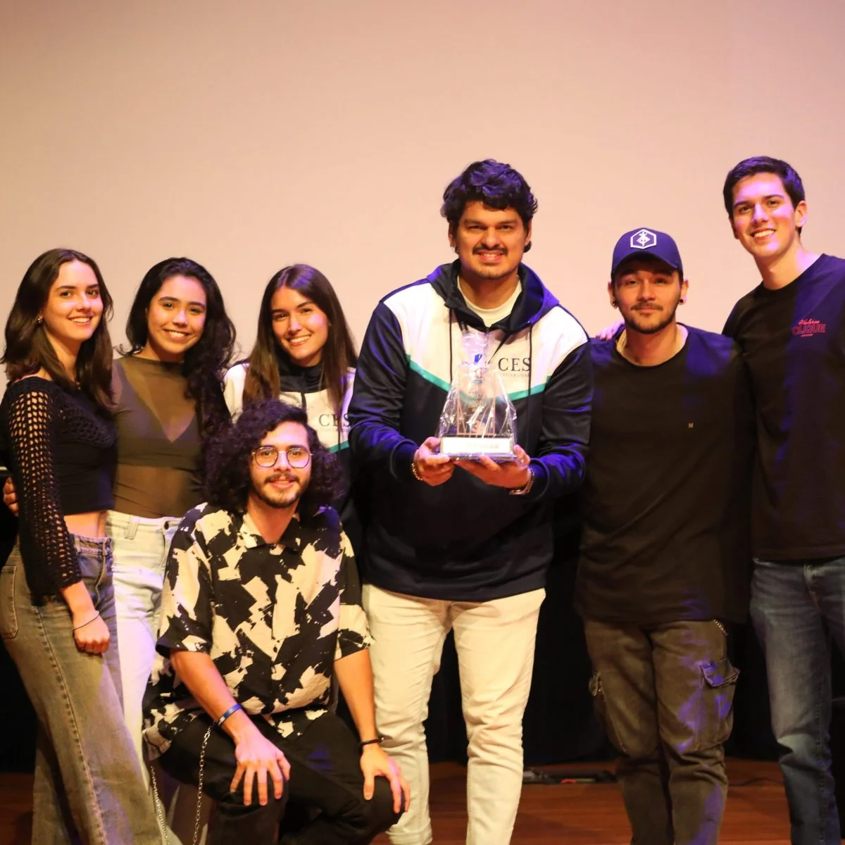 Grupo de varias personas posando juntas en un escenario; una persona al centro sostiene un trofeo transparente. Todas miran a la cámara, con iluminación cálida y fondo liso.