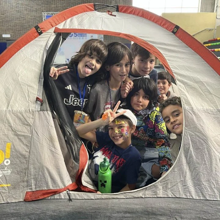 Grupo de niños dentro de una tienda de campaña, algunos con credenciales colgadas y uno con el rostro pintado, en un espacio interior.