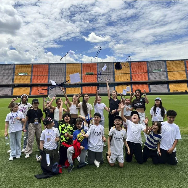 Grupo de niños en un campo deportivo, levantando y lanzando objetos al aire frente a las gradas de un estadio.