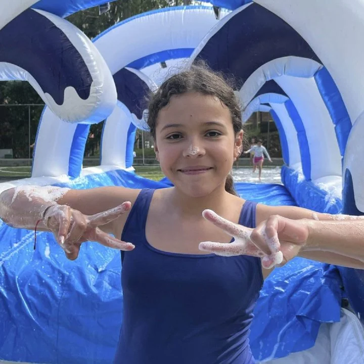 Persona niña frente a un inflable con agua y espuma, levantando ambas manos en forma de señal de paz, en un entorno exterior.