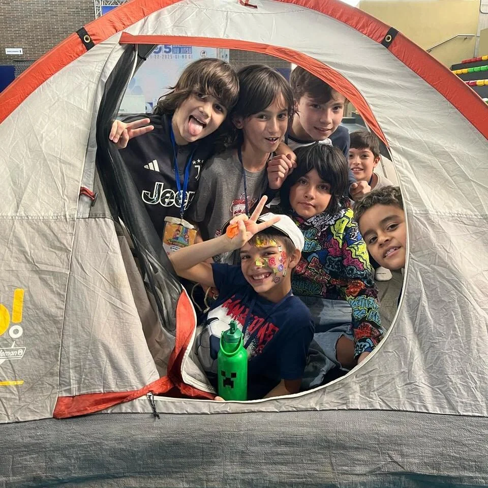 Grupo de siete niños reunidos dentro de una carpa gris y naranja, mirando hacia la cámara; uno de los niños tiene pinturas en la cara y sostiene una botella verde.