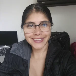 Persona con gafas de montura clara, vistiendo una chaqueta oscura, frente a un escritorio con un computador portátil en un entorno interior.
