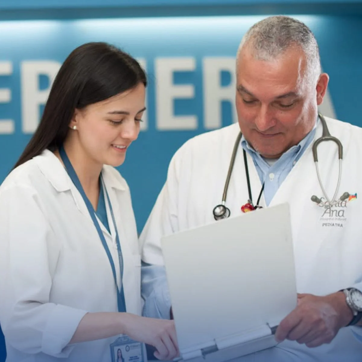 Dos profesionales de la salud con batas blancas revisan juntos un documento en un entorno clínico, con un estetoscopio visible y fondo azul institucional.