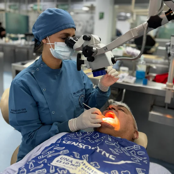Persona con indumentaria clínica utiliza un microscopio mientras realiza un procedimiento odontológico a otra persona reclinada, bajo iluminación especializada en un consultorio.