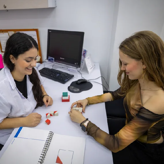 Dos personas en un consultorio realizan una actividad con figuras y materiales didácticos sobre un escritorio, junto a un computador y cuadernos de trabajo.