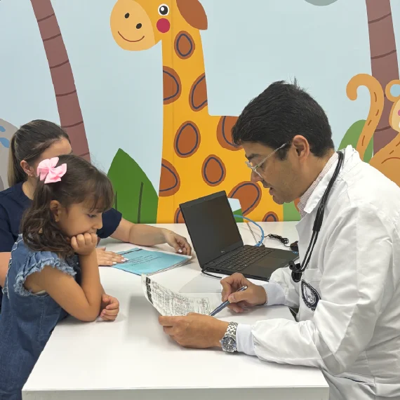 Profesional de la salud revisa documentos con una persona menor de edad y otra persona acompañante en un consultorio, con computador portátil y decoraciones infantiles en la pared.