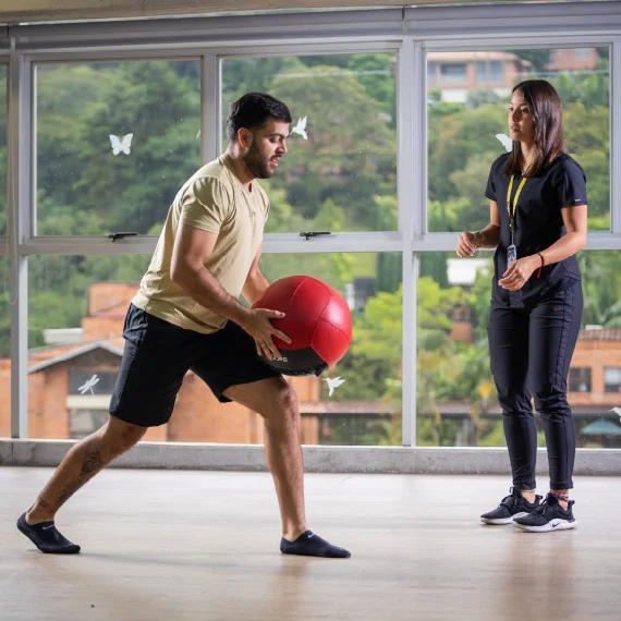 Dos personas realizan un ejercicio físico con una pelota grande en un espacio interior, mientras una supervisa el movimiento frente a ventanas con vista exterior.