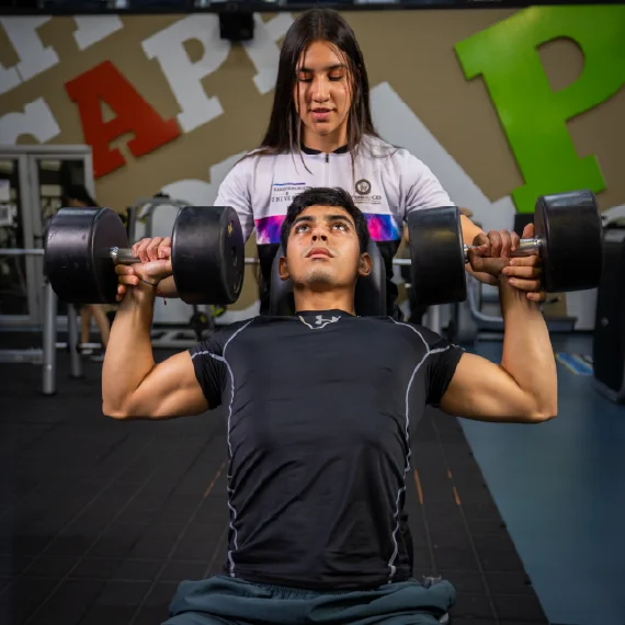 Dos personas realizan un ejercicio de fuerza con pesas en un gimnasio, mientras una supervisa la técnica durante el levantamiento en un entorno con equipos de entrenamiento.