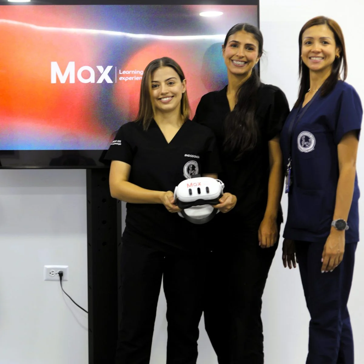 Tres personas con uniforme clínico posan frente a una pantalla con el texto “MaX Learning experience”; una de ellas sostiene un visor de realidad virtual.