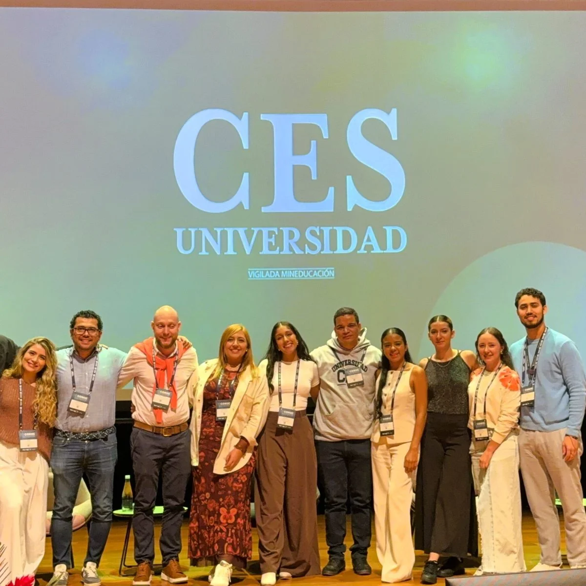 Grupo de varias personas con credenciales posan en un escenario frente a una pantalla con el logo “CES Universidad” y el texto “Vigilada Mineducación”, durante un evento académico.