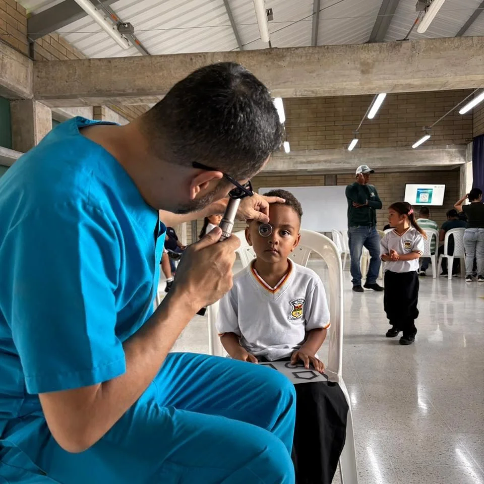 Profesional de salud realiza un examen visual a un niño sentado en una silla, utilizando un instrumento oftalmológico, mientras otras personas esperan en un espacio comunitario.