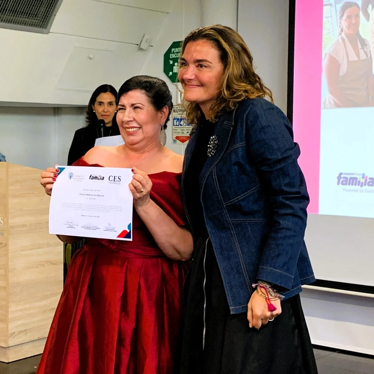 Persona en un auditorio recibe un certificado de la Universidad CES y lo sostiene mientras posa junto a otra persona; al fondo se observa una presentación proyectada y una tercera persona con micrófono.