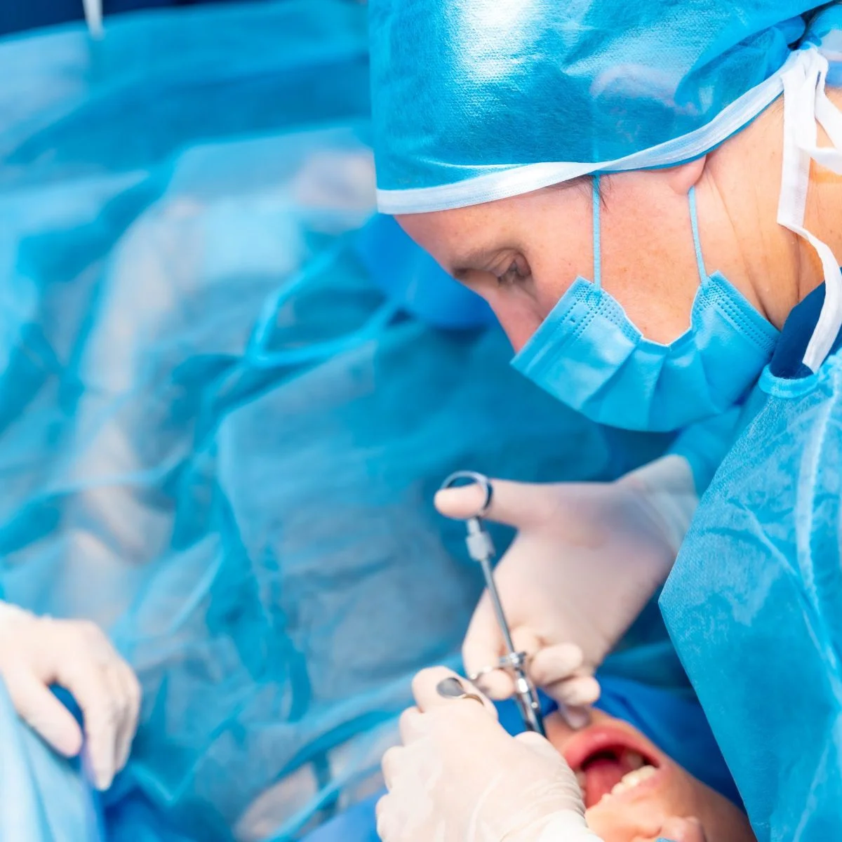 Profesional de salud con equipo quirúrgico realiza un procedimiento dental utilizando instrumentos dentro de la boca de un paciente, en un entorno clínico.
