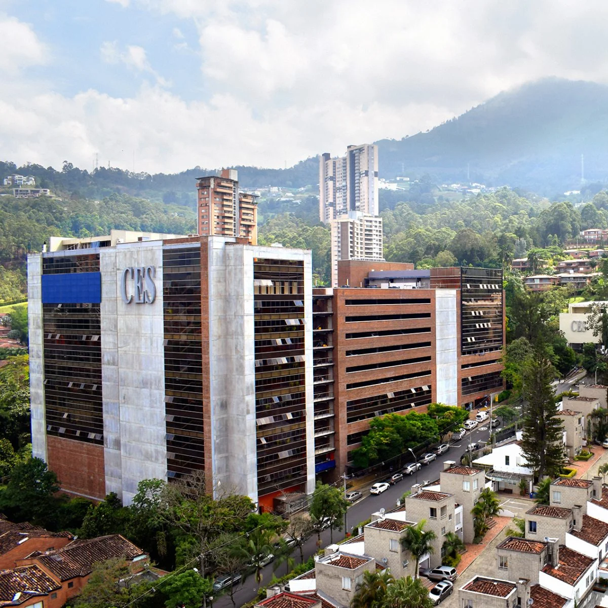 Vista urbana de un campus universitario con edificios modernos, incluido uno con el letrero “CES”, rodeado de viviendas, árboles y montañas bajo un cielo nublado.
