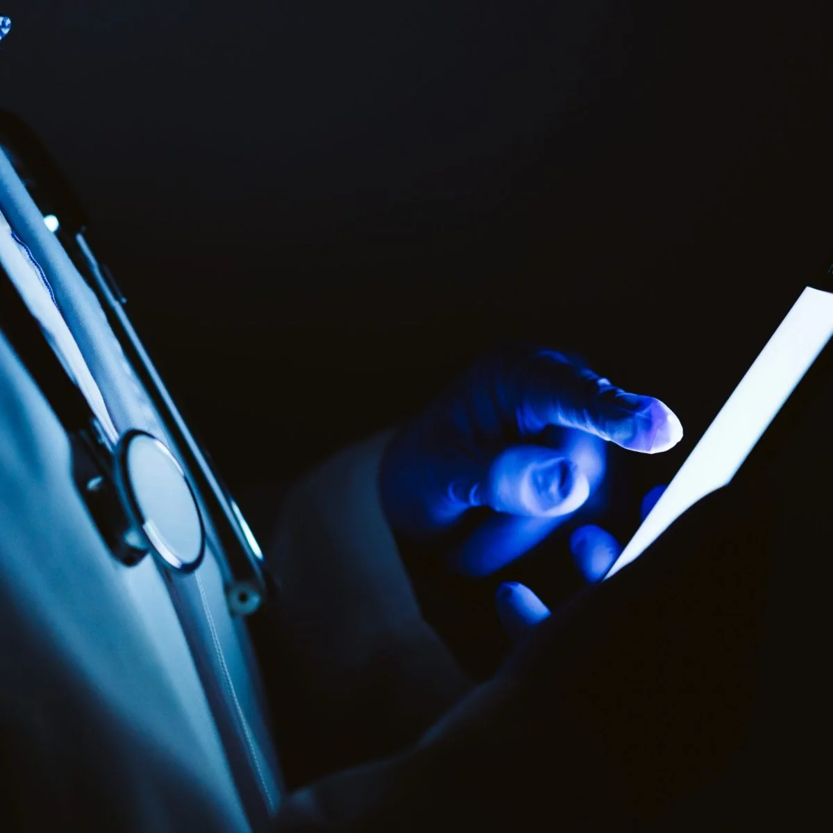Profesional de la salud con estetoscopio usando un teléfono móvil en un entorno oscuro con iluminación azul.