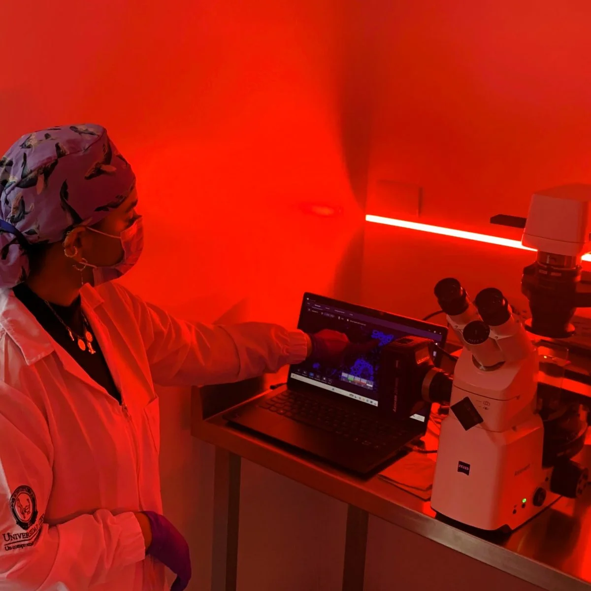 Persona en laboratorio iluminado con luz roja manipula un equipo junto a un microscopio y un computador portátil.