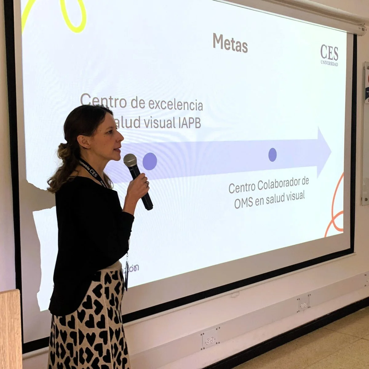 Persona sostiene un micrófono y presenta frente a una pantalla con una diapositiva titulada “Metas” de la Universidad CES, sobre un centro de excelencia en salud visual.