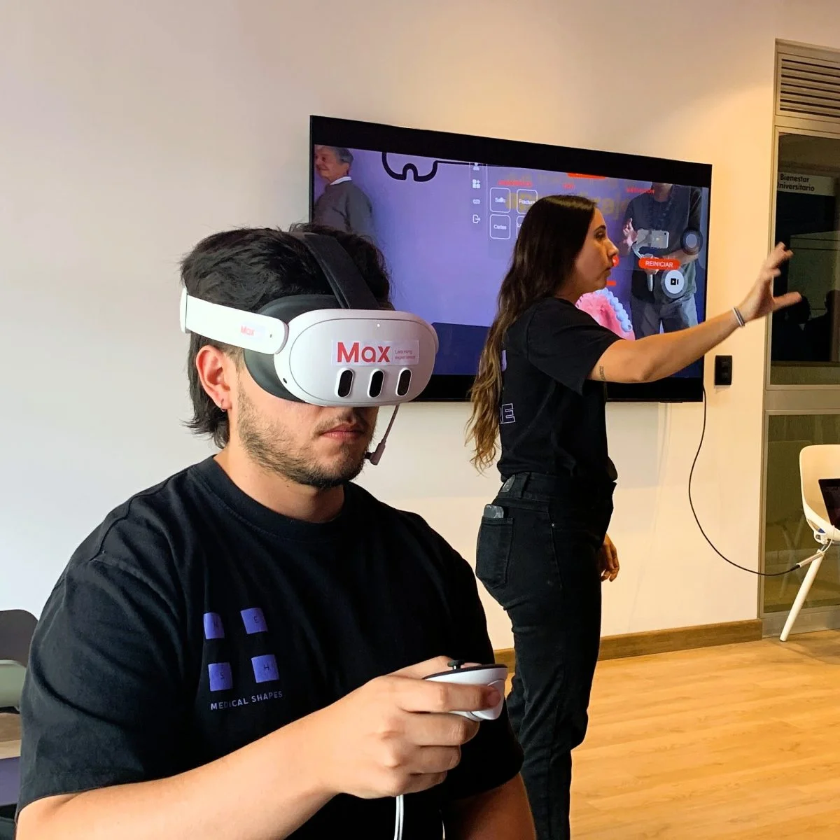 Persona utilizando un visor de realidad virtual y sosteniendo un controlador mientras participa en una experiencia inmersiva en un espacio interior. Al fondo, otra persona interactúa con una pantalla que muestra el contenido de la simulación.