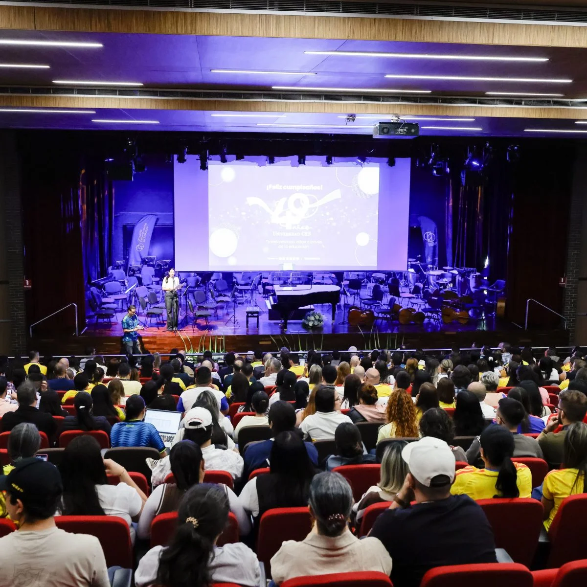Vista general de un auditorio con gran afluencia de público. En el escenario se observan instrumentos de orquesta, un piano de cola y una pantalla de proyección iluminada, en el marco de un evento institucional de la Universidad CES.