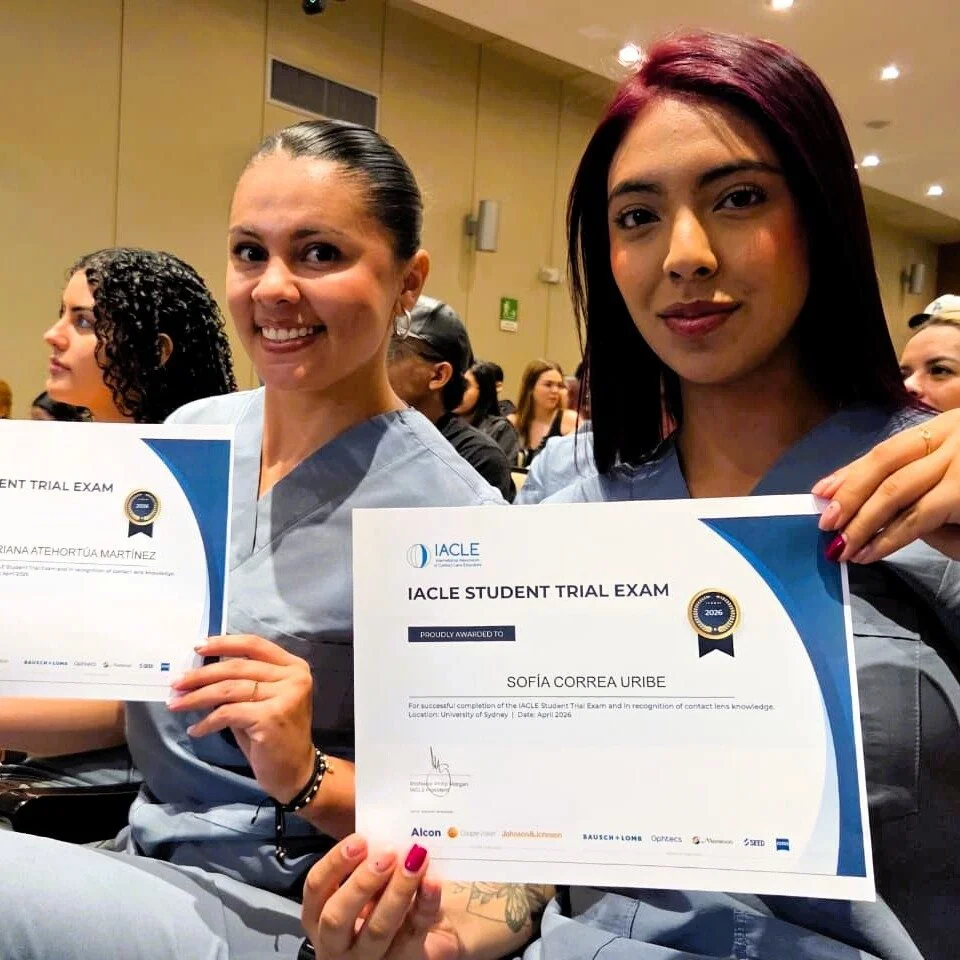 Dos personas con uniforme clínico azul claro sostienen certificados del examen IACLE Student Trial Exam durante un evento académico realizado en un auditorio. En segundo plano se observan otros asistentes sentados.