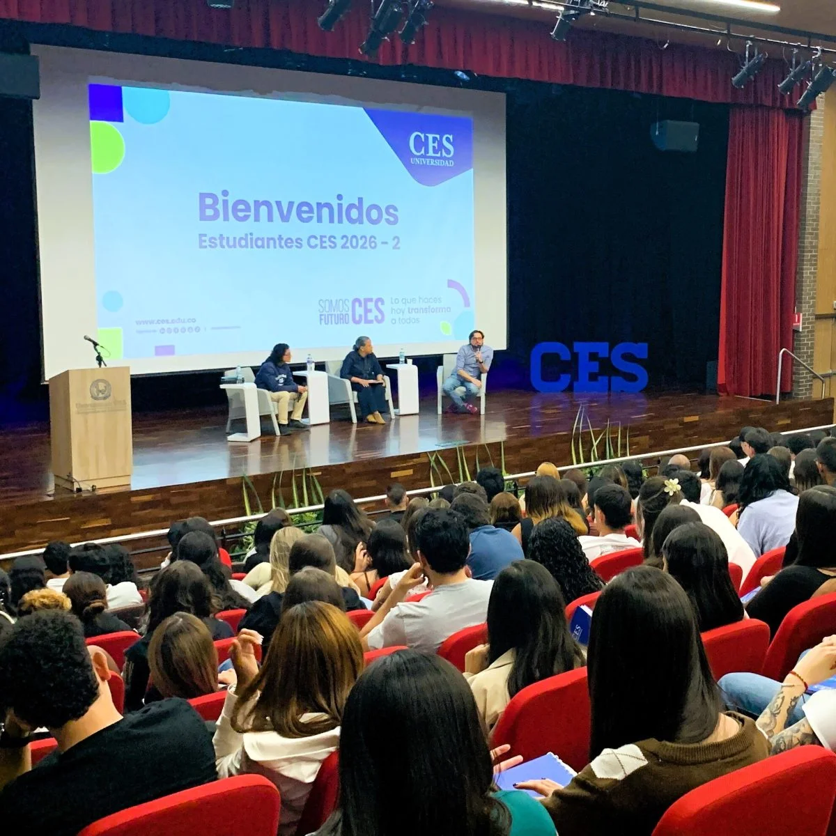 Auditorio universitario con estudiantes asistentes a una jornada de bienvenida. En el escenario, tres panelistas participan en una conversación frente al público, junto a una pantalla que proyecta el mensaje “Bienvenidos Estudiantes CES 2026-2” y el logo de la Universidad CES.