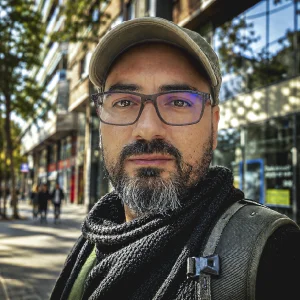 Persona al aire libre vistiendo gafas, gorra y bufanda oscura, con una mochila al hombro, en una calle urbana rodeada de edificios y árboles.