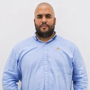 Persona de pie frente a un fondo liso claro, vistiendo una camisa de botones azul claro de manga larga.