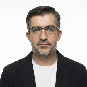 Persona de pie frente a un fondo liso claro, vistiendo una chaqueta oscura sobre una camiseta blanca y gafas de montura rectangular.