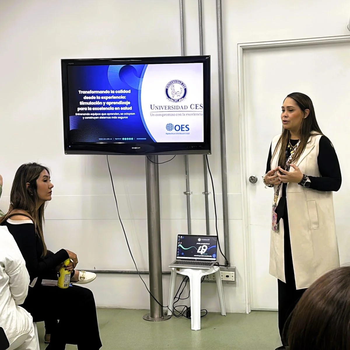 Persona realizando una presentación en un aula o sala de capacitación frente a un grupo de asistentes. En una pantalla se muestra una diapositiva de la Universidad CES sobre simulación y aprendizaje para la excelencia en salud. Debajo de la pantalla hay un computador portátil conectado al sistema de presentación, mientras varias personas observan la exposición.