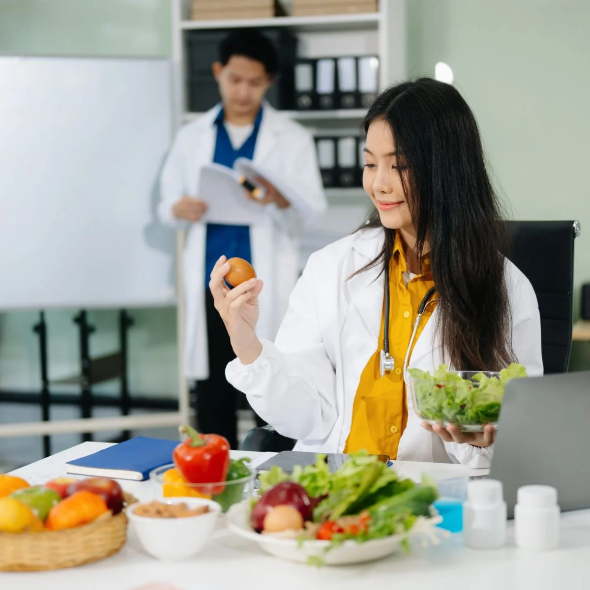 Profesional de la salud en un consultorio observa un alimento mientras sostiene un recipiente con vegetales. Sobre el escritorio hay frutas, verduras, suplementos nutricionales y un computador portátil, en un entorno relacionado con la educación alimentaria y la nutrición.