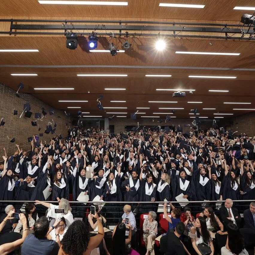 Grupo numeroso de personas con toga y estola durante una ceremonia de graduación en un auditorio. Varias personas lanzan sus birretes al aire mientras asistentes observan y toman fotografías desde la parte inferior de la imagen.