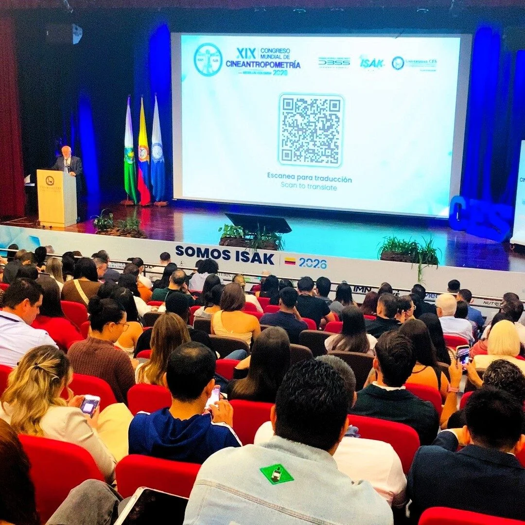 Auditorio con público asistente observando una presentación en escenario durante el XIX Congreso Mundial de Cineantropometría ISAK 2026. En la pantalla principal se muestra un código QR para traducción del evento, junto a los logotipos de las organizaciones participantes y varias banderas ubicadas al lado del podio.