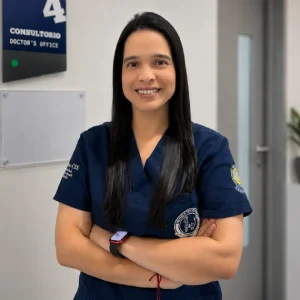 Persona de pie con los brazos cruzados, vistiendo uniforme clínico azul oscuro con emblemas institucionales visibles, en un pasillo de atención médica junto a la señalización de un consultorio.