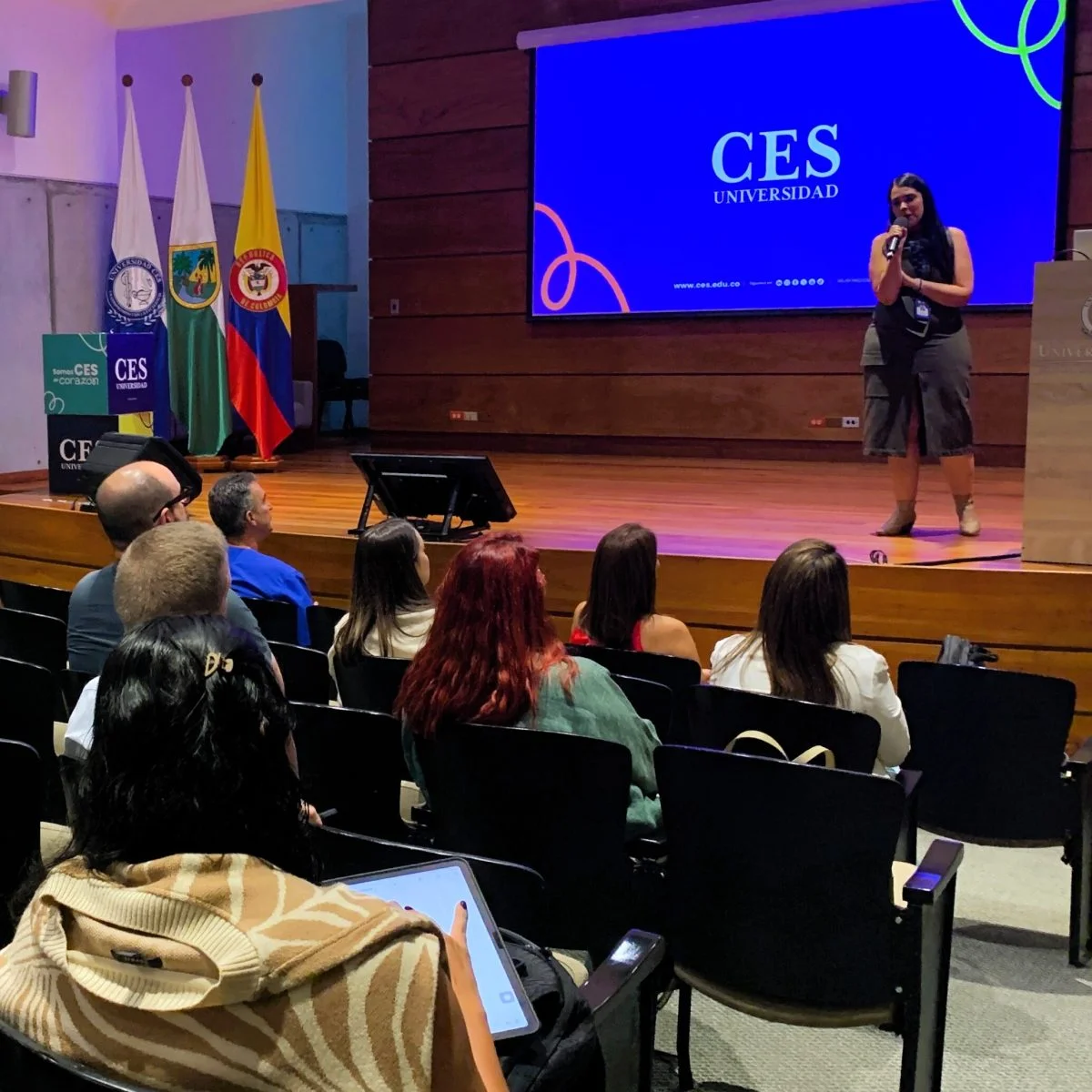 Fotografía de un evento académico realizado en un auditorio de la Universidad CES. En el escenario, una persona realiza una presentación con micrófono frente a una audiencia sentada. Detrás se observa una pantalla con el logo de la Universidad CES y, a un costado, varias banderas institucionales y de Colombia. La imagen representa una actividad de divulgación, formación o intercambio de conocimientos en un entorno universitario.