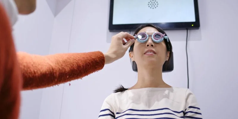 Persona sentada en una clínica oftalmológica durante un examen visual. Un profesional ajusta unas gafas de prueba mientras una pantalla de evaluación se observa al fondo.