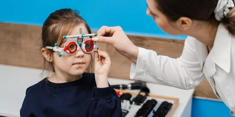 Niña durante un examen visual en un consultorio oftalmológico. Un profesional de la salud ajusta unas gafas de prueba mientras realiza la evaluación de la visión.