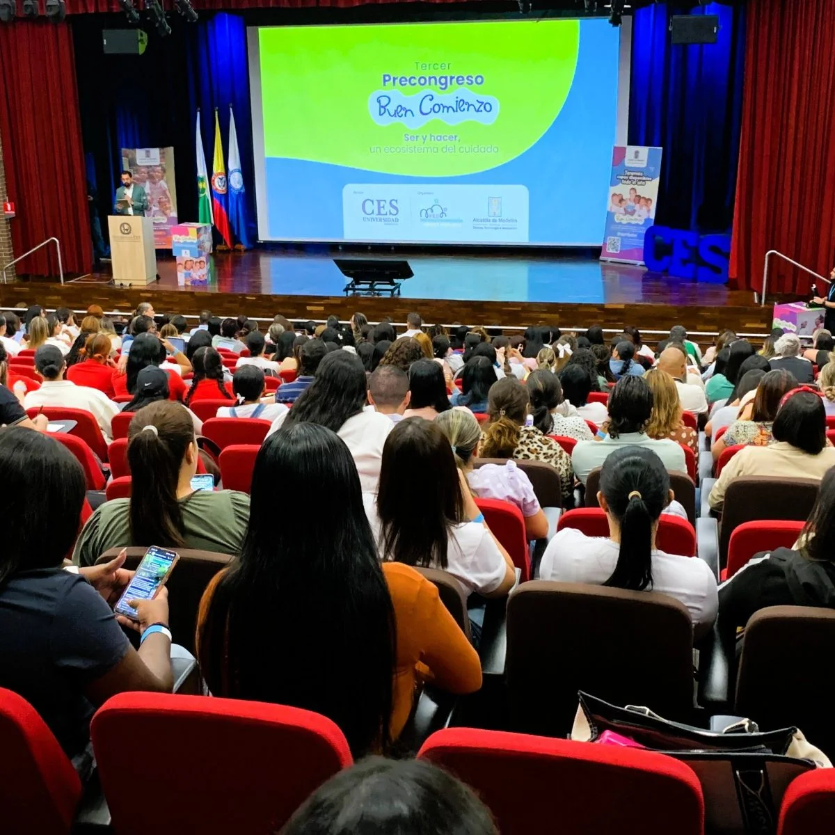 Auditorio con gran asistencia de público sentado frente a un escenario. En la pantalla principal se proyecta la presentación del evento “Tercer Precongreso Buen Comienzo: Ser y hacer, un ecosistema del cuidado”, con logos institucionales de la Universidad CES, Buen Comienzo y la Alcaldía de Medellín. A un lado del escenario hay una persona realizando una intervención desde un atril, junto a banderas y material promocional del evento.