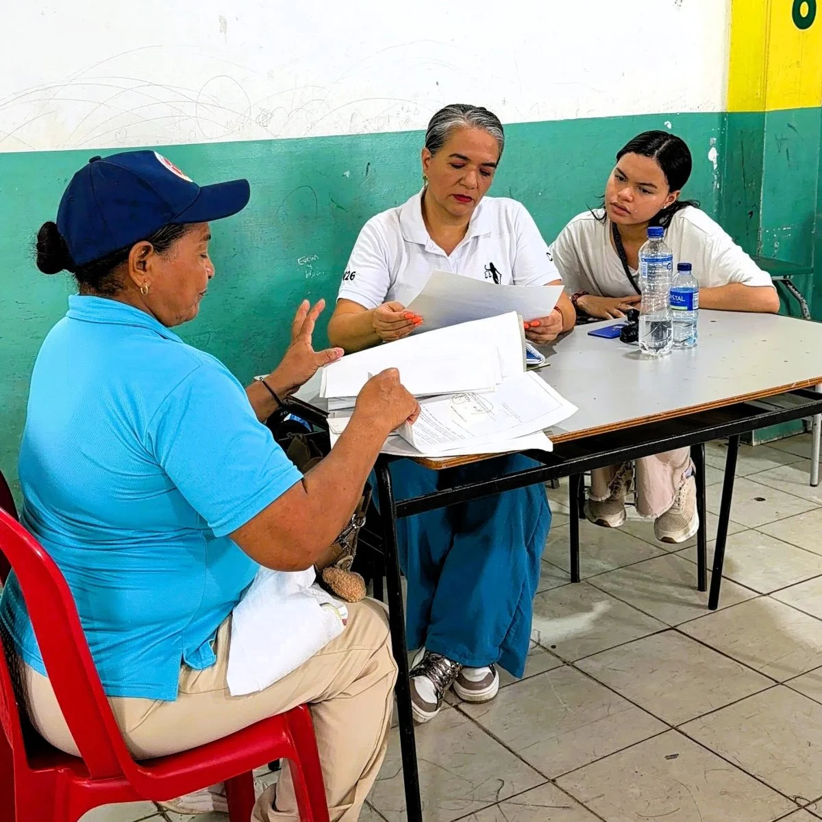 Tres personas están reunidas alrededor de una mesa en un salón. Una persona sentada en una silla roja muestra varios documentos mientras otras dos revisan hojas impresas. Sobre la mesa hay papeles, un teléfono móvil y dos botellas de agua. El espacio tiene paredes blancas y verdes y piso de baldosas.